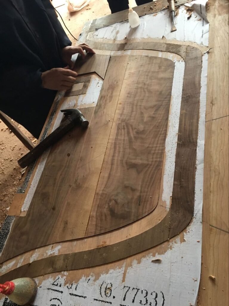 An artisan's hands carefully fitting solid wood panels onto a custom door template layout on a workshop table, alongside a hammer and wood glue during the fabrication process.