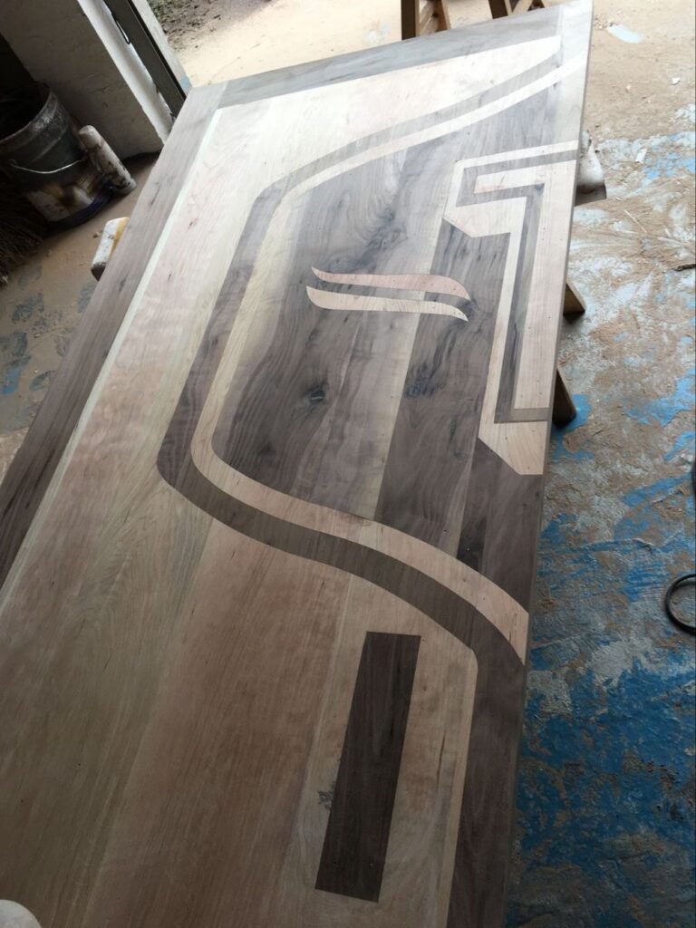 An unfinished custom wood door panel lying on a workbench in a woodworking shop, showcasing raw black walnut and cherry wood inlay details forming a geometric shield and 'T' emblem before the final finish.