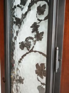 Close-up angled view of a heavy-duty steel hinge mounted on the dark operable wrought iron frame of a custom solid wood entry door with textured privacy glass.