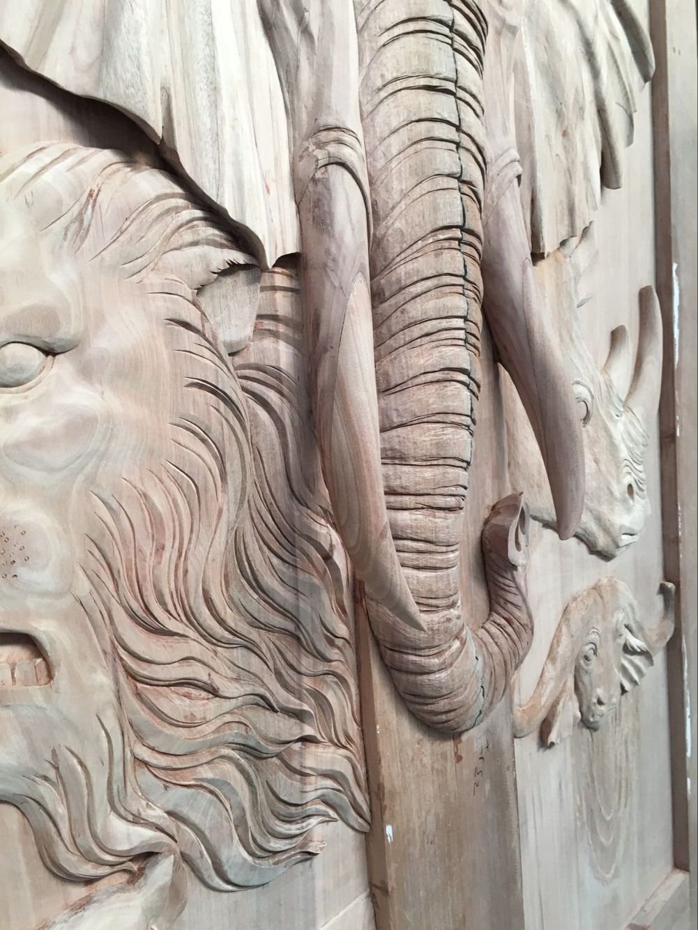 A detailed close-up side angle of a raw, unfinished solid wood door panel showcasing intricate, high-relief hand carvings of an African elephant trunk, long tusks, a textured lion mane, a rhinoceros, and a cape buffalo.