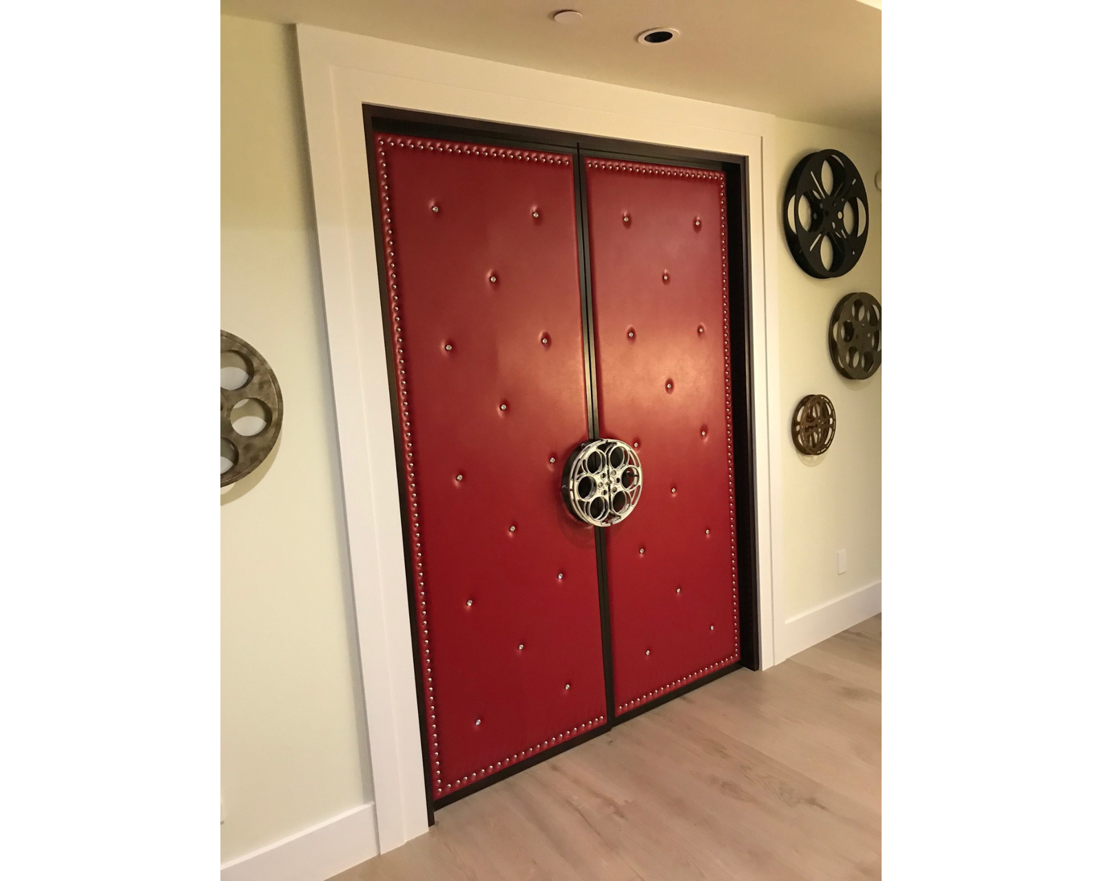 A fully installed custom red upholstered acoustic double door for a home theater by Crown Custom Doors, featuring diamond-tufted vinyl with crystal studs, silver nailhead trim, and a custom silver film reel handle, set in a modern hallway decorated with vintage film reel wall art.