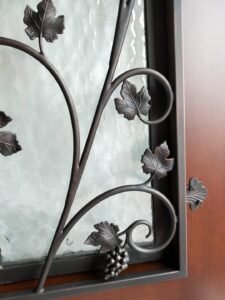 Side-angle close-up of a custom front door showing the precise fitment of a hand-forged wrought iron grapevine grille and textured glass insert into a finished solid wood frame.