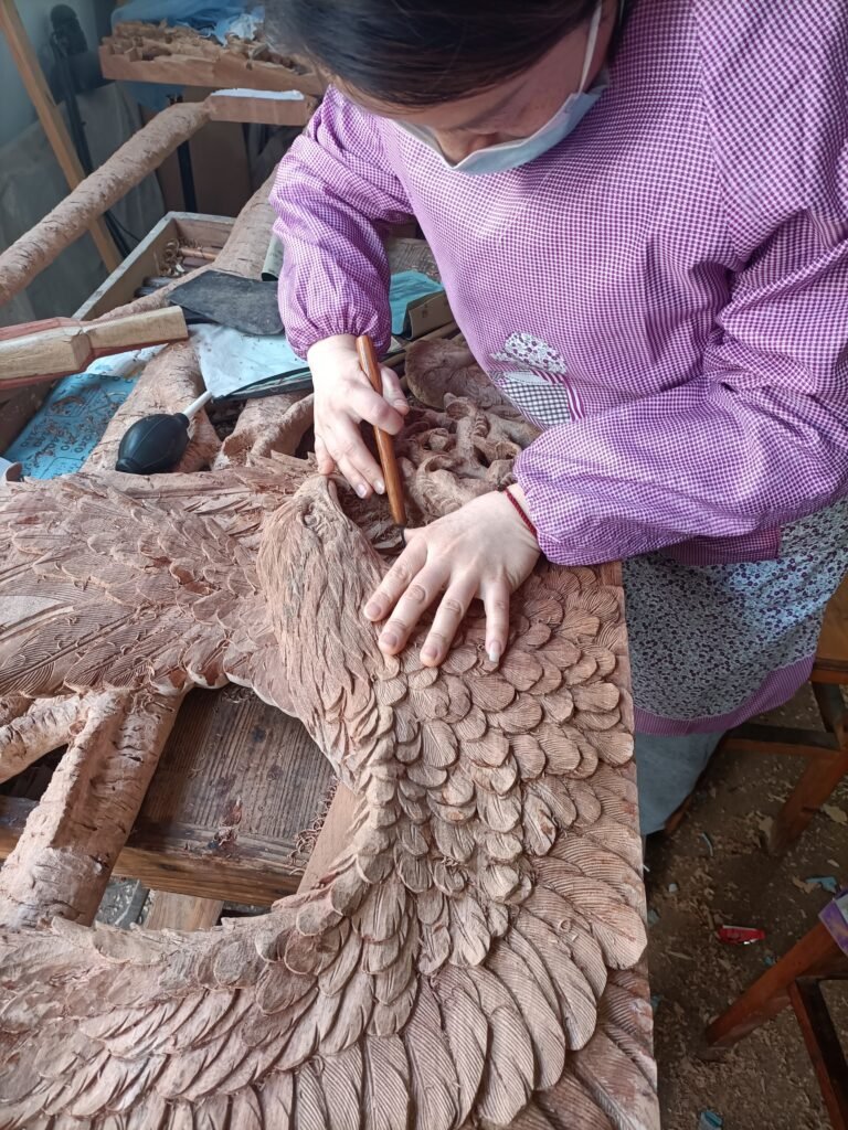 Master artisan using hand gouge chisels to carve fine feather details on a solid wood eagle door panel in a woodworking workshop.