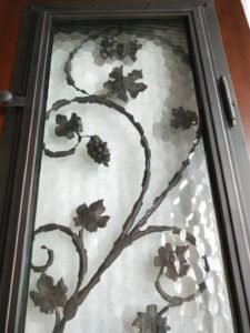 Low-angle view of an operable wrought iron grapevine insert featuring a latch mechanism and textured privacy glass inside a custom exterior door frame.