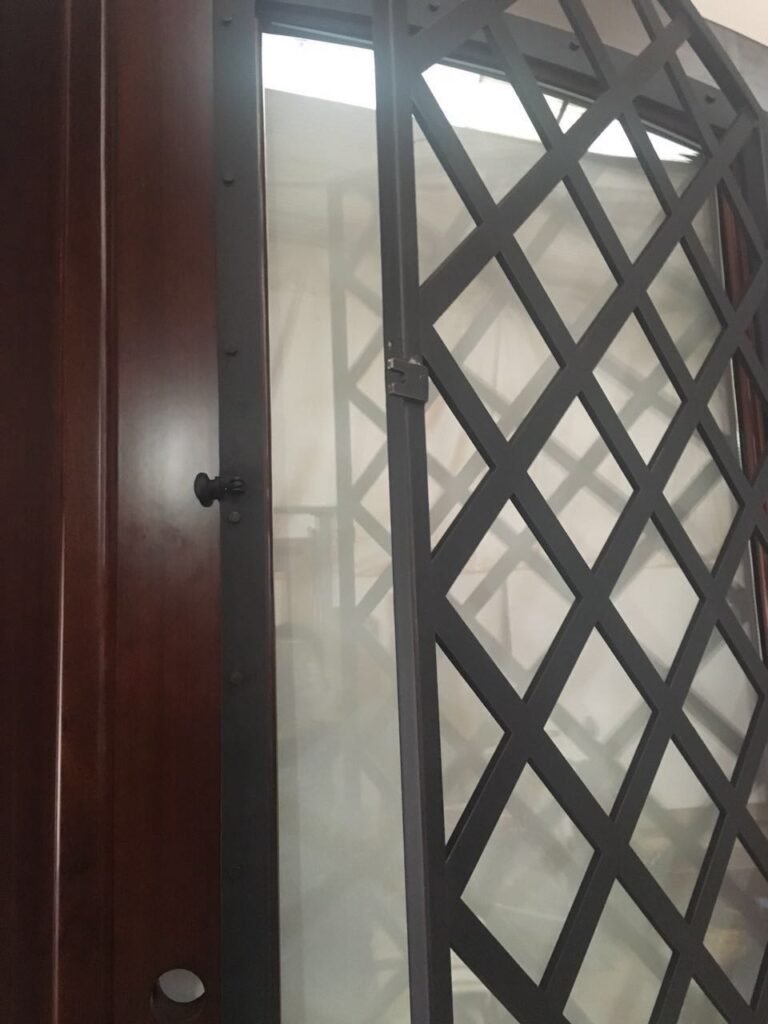 A functional detail image of a custom door by Crown Custom Doors, showing the black geometric lattice iron grille panel unlatched and swung open on its hinges, exposing the dual-pane glass insert and the rich mahogany wood frame behind it.