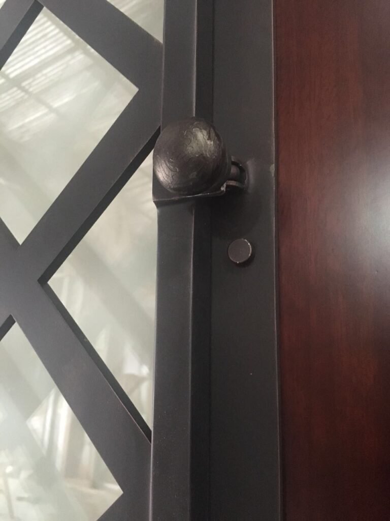 A macro detail shot of a custom door by Crown Custom Doors, showcasing a dark oil-rubbed bronze cast iron knob and latch mechanism attached to an operational geometric lattice iron grille panel, mounted over a rich mahogany wood entry frame.