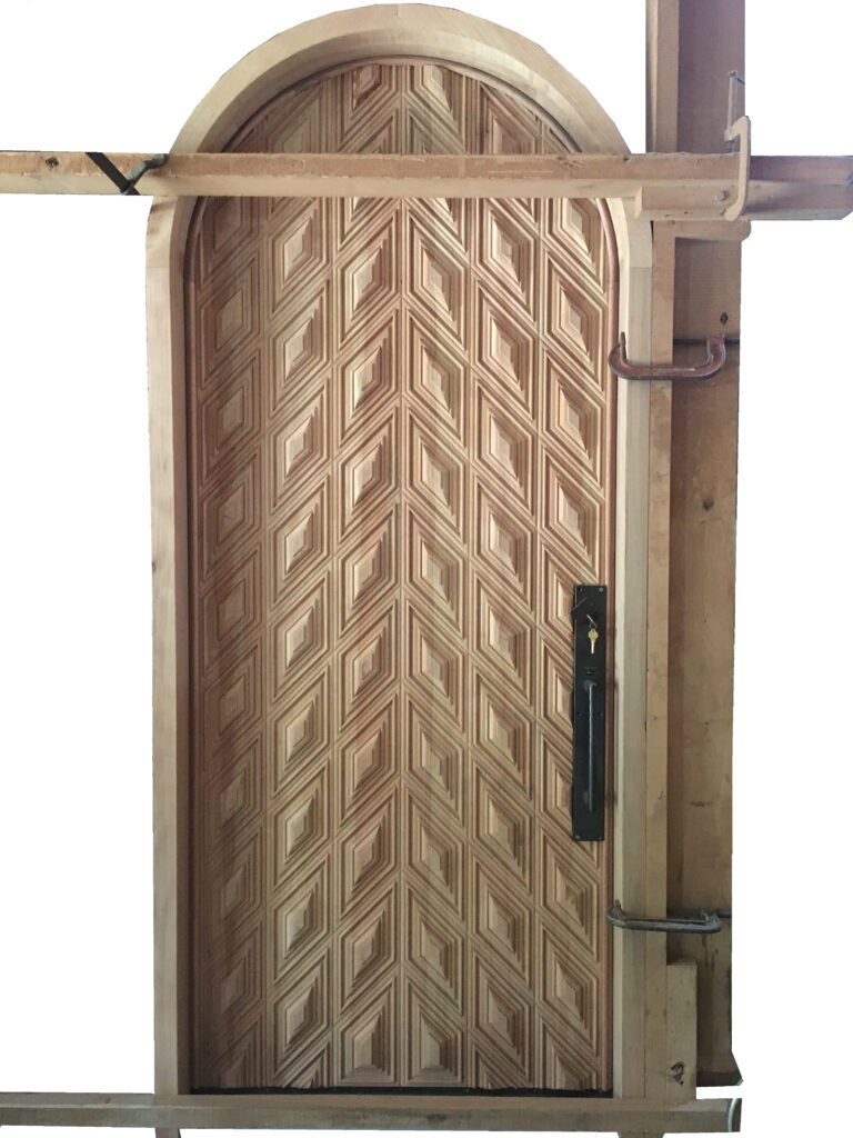 An unfinished raw wood custom arched single entry door by Crown Custom Doors during the pre-hung assembly and testing process in the workshop, showcasing the intricate diamond chevron parquet panels mounted inside its matching rounded frame casing with temporary wood braces and hardware.