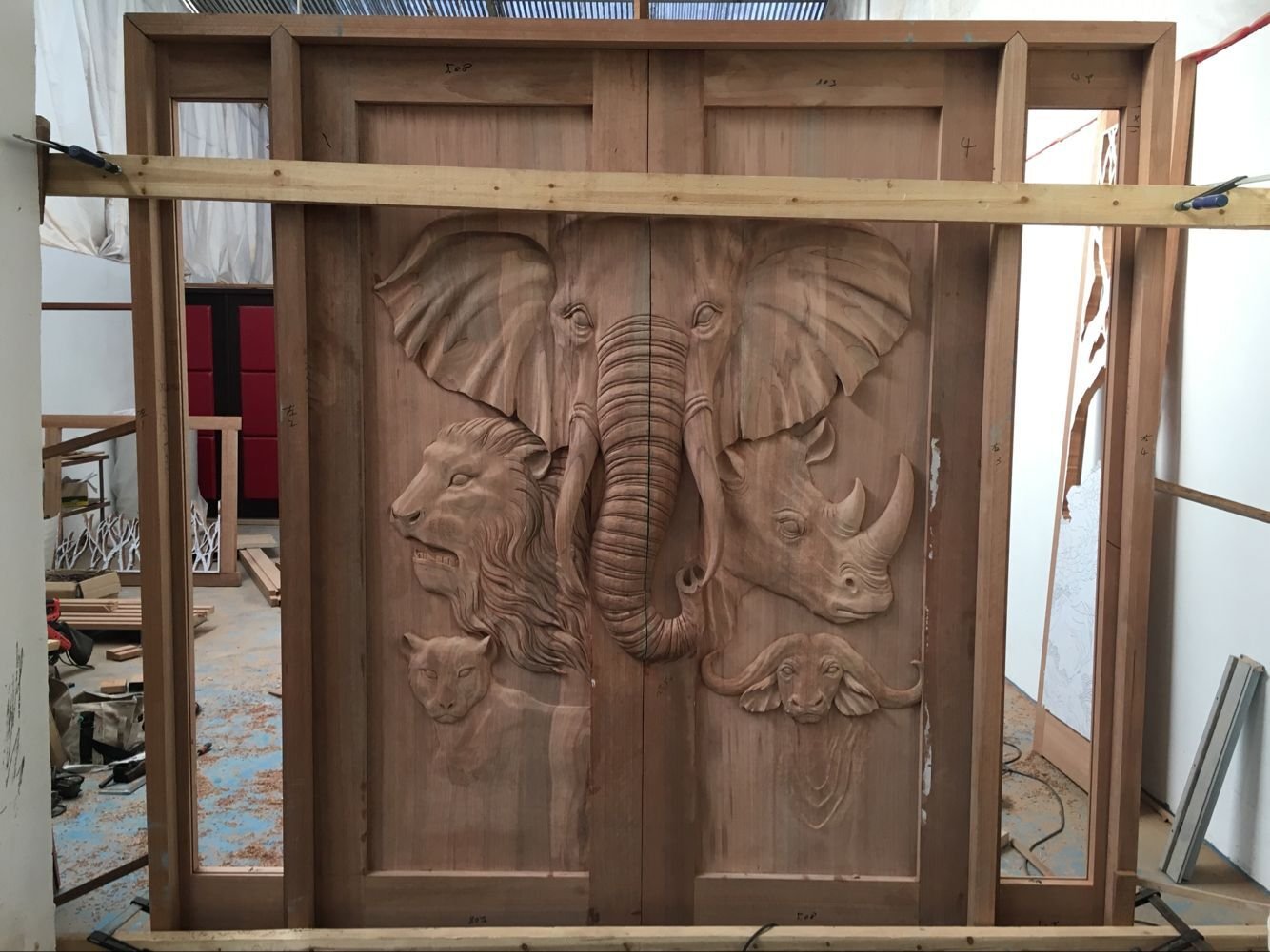 A raw, unfinished solid wood double entry door featuring high-relief hand carvings of an elephant, lion, rhinoceros, leopard, and cape buffalo, shown clamped inside a wooden framing template in a custom woodworking workshop.