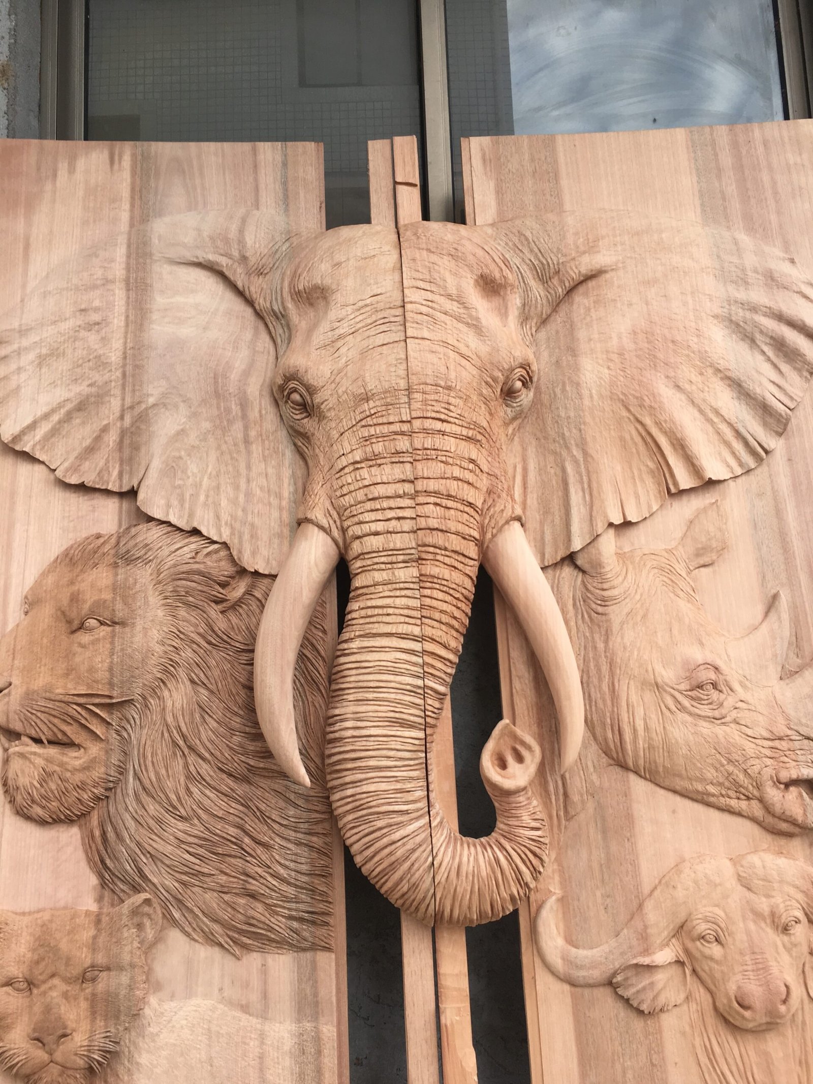 A detailed front view of a raw, unfinished solid wood double entry door panel featuring highly detailed, high-relief hand carvings of a prominent African elephant head, flanked by a lion, rhinoceros, leopard, and cape buffalo in a workshop.