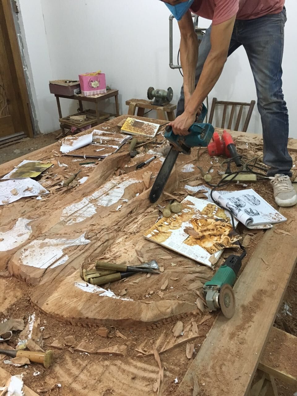 A master artisan in a woodworking workshop standing over a massive solid wood slab, using a power carver chainsaw to rough out the high-relief shapes of the Wild Africa door amidst wood shavings, hand chisels, and reference drawings.