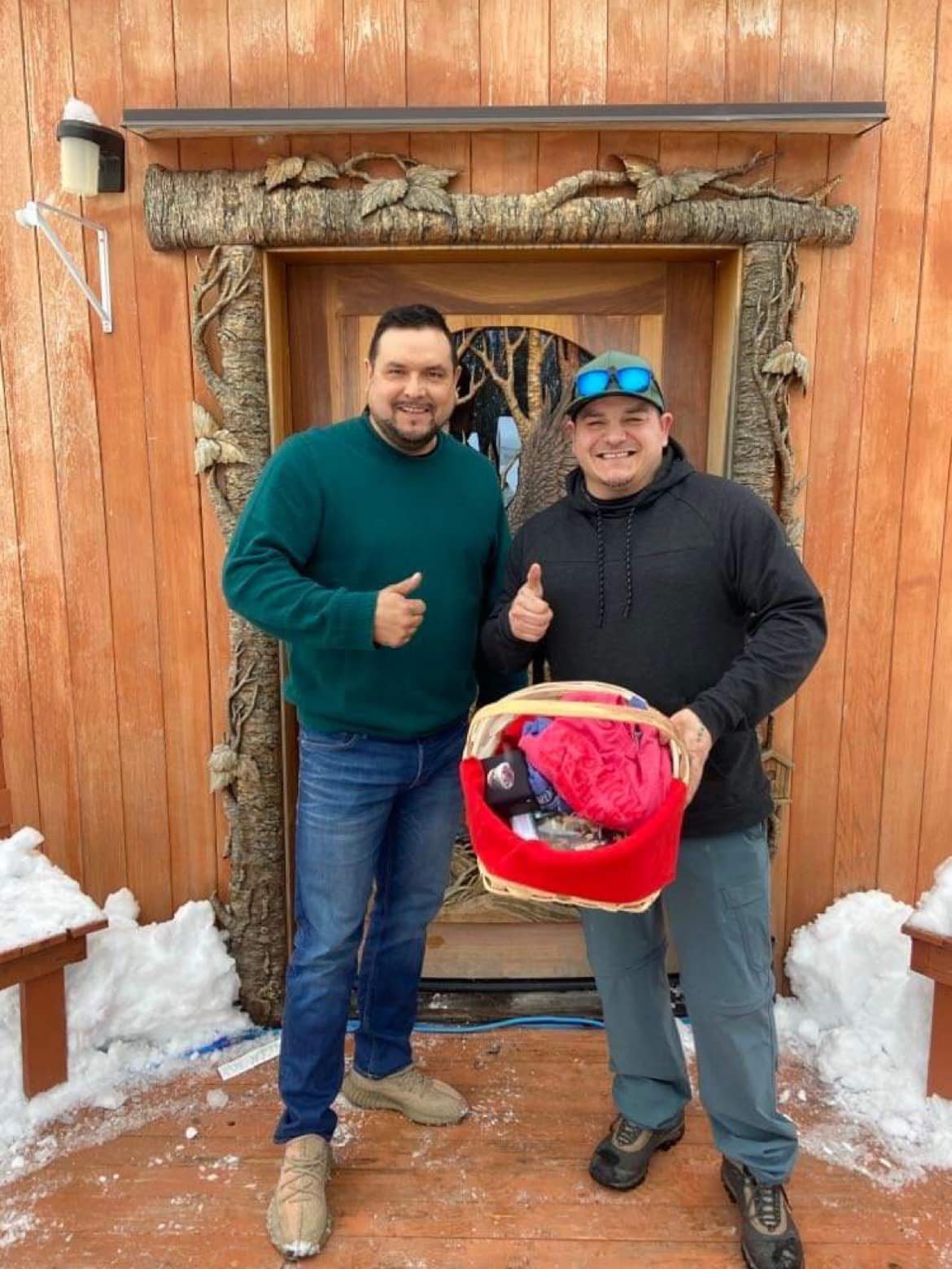 Two happy male clients giving a thumbs up in front of an installed custom hand-carved mahogany bald eagle front door with sculpted tree casing on a wooden yurt in snowy winter weather.