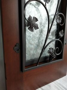 A close-up side angle of a premium solid wood door by Crown Custom Doors, showing a dark brown stained timber panel integrated with a hand-forged wrought iron grapevine scrollwork grille, detailed grape clusters, and decorative leaf hinges over textured frosted privacy glass.