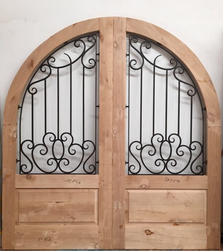 Handcrafted full-arched double doors made of unfinished solid pine wood by Crown Custom Doors, featuring intricate black wrought iron scrollwork grilles integrated into the upper glass panels within a workshop setting.