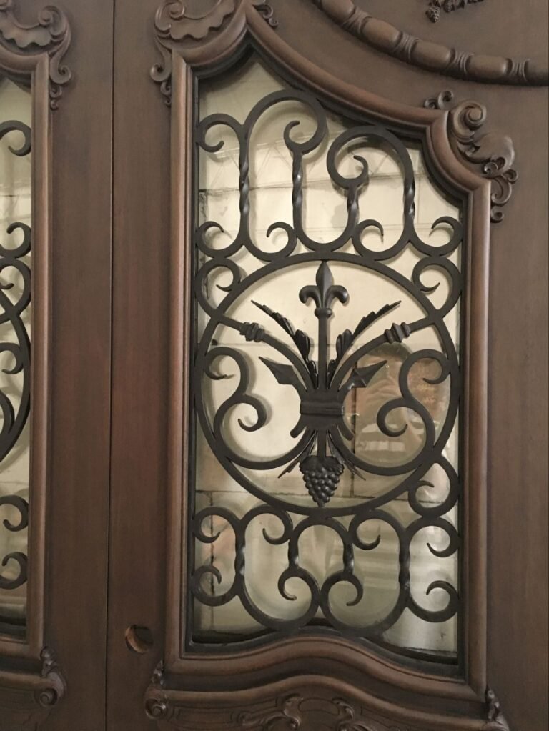 Close-up detail of a custom solid wood door featuring a hand-forged black wrought iron insert with a central Fleur-de-lis and grape cluster motif, surrounded by elegant, hand-carved baroque wood trim.