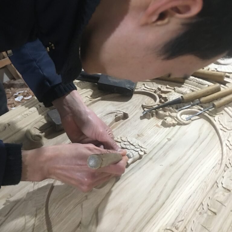 Close-up angle of a craftsman's hands using a fine palm gouge chisel to carefully sculpt delicate hair and feather textures on a custom solid wood door panel at Crown Custom Doors.