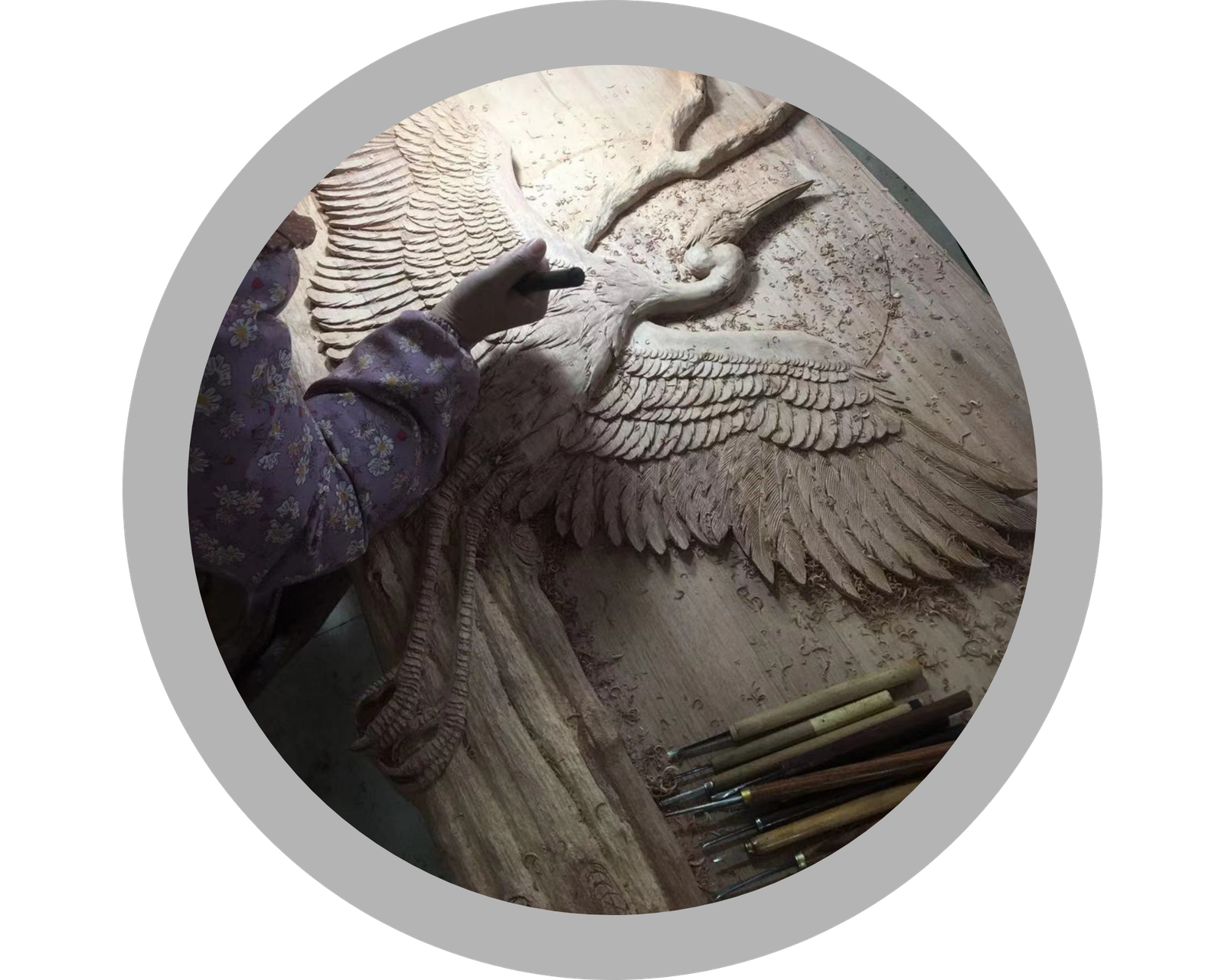 Overhead view of an artisan hand-chiseling intricate feather details into a solid wood heron carving on a workshop table surrounded by woodworking tools and wood shavings.