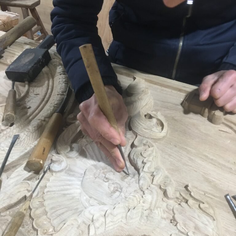 Close-up action shot of a master craftsman hand-chiseling intricate scrollwork and face details on a custom solid wood door panel using traditional woodworking chisels and mallets at Crown Custom Doors.