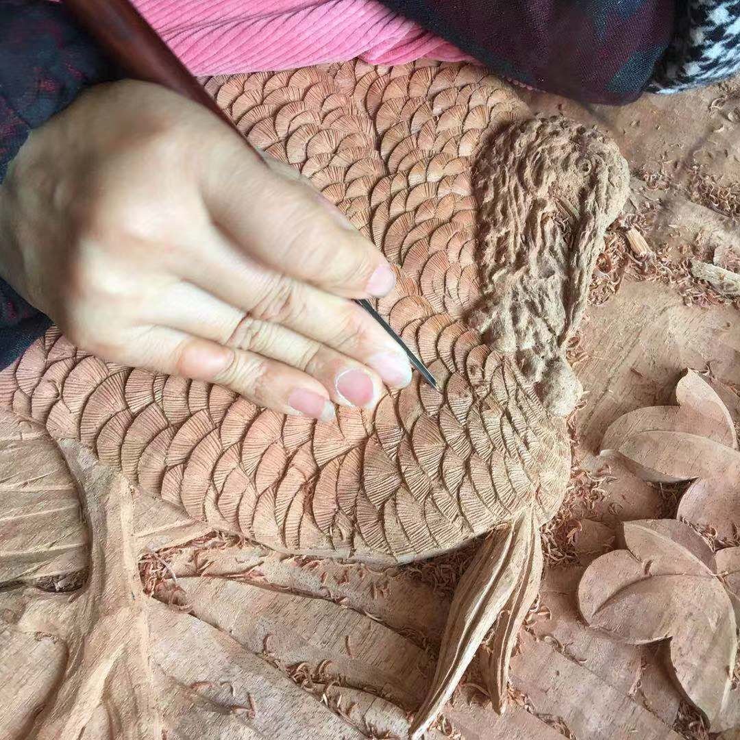 Close-up, top-down view of a woodworker's hand using a small chisel to carve delicate, overlapping feather textures into a raw solid wood relief sculpture of a wild turkey, surrounded by wood shavings.