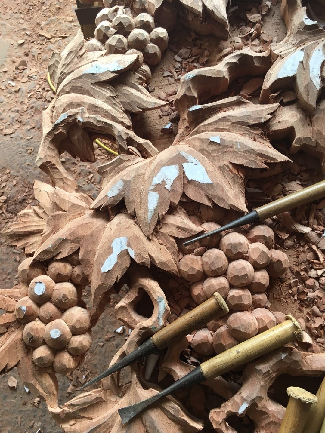 Close-up view of traditional woodworking chisels lying across raw, hand-carved solid wood grapevine leaves and grape clusters surrounded by wood shavings in a workshop.