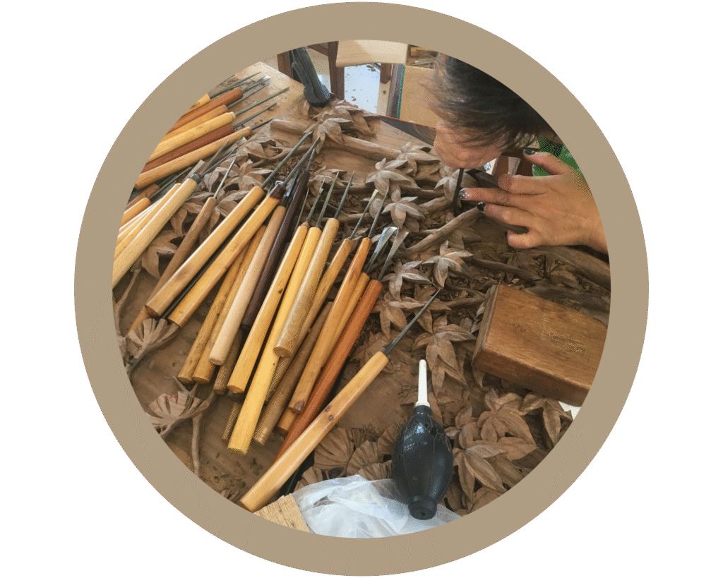 Circular overhead view of an artisan's hands carefully carving detailed leaf patterns on a solid wood panel using specialized chisels and traditional woodworking tools in a workshop.