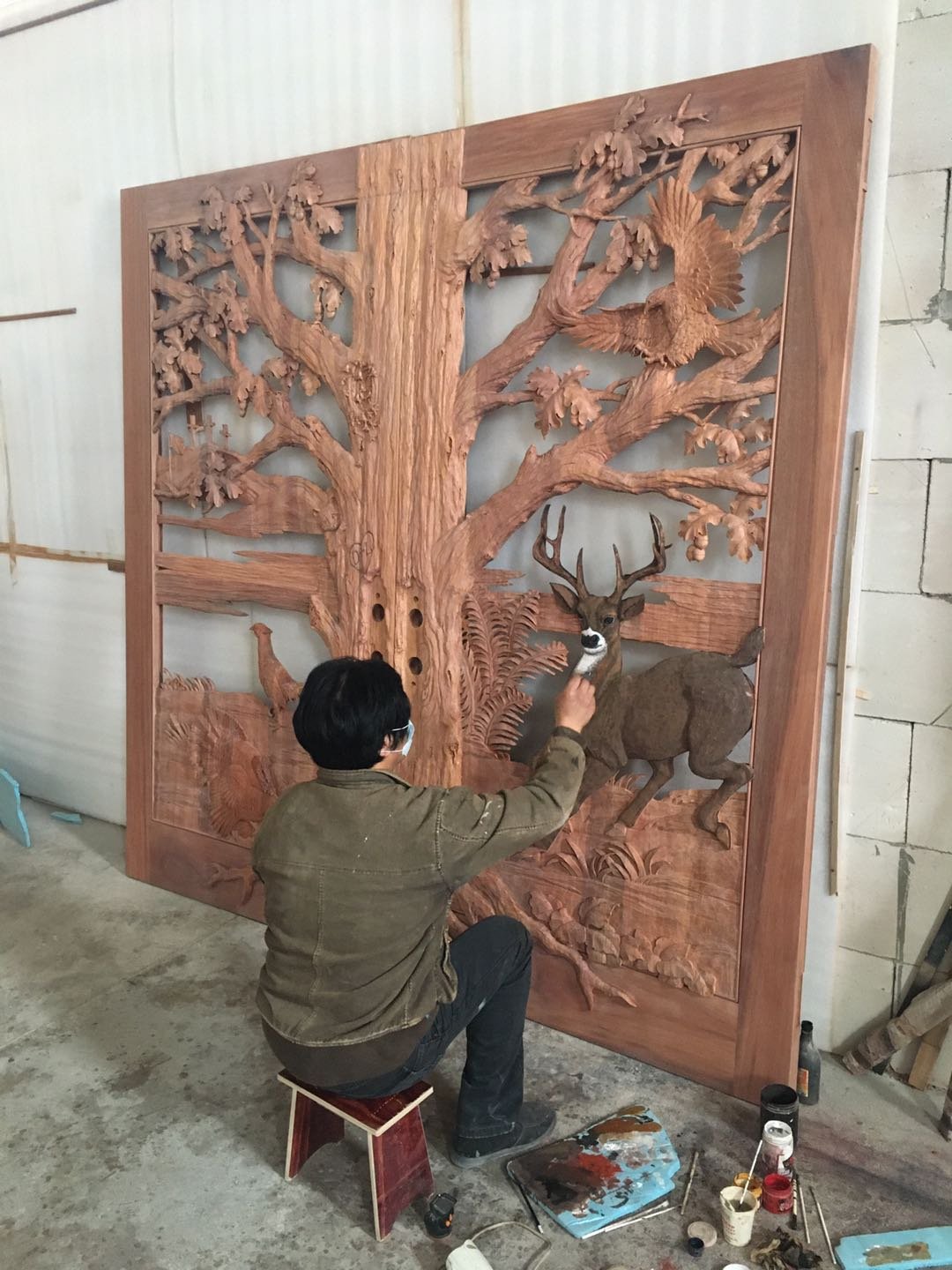 Workshop photograph of an artisan craftsman seated on a small bench, carefully hand-painting facial highlights on a high-relief carved white-tailed deer buck across large solid wood double entry doors featuring an oak tree canopy design.
