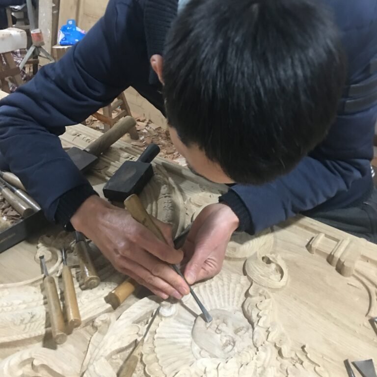 Overhead shot of a master woodcarver intently focused on hand-chiseling fine relief facial features and ornamental details on a raw solid wood door panel at Crown Custom Doors.