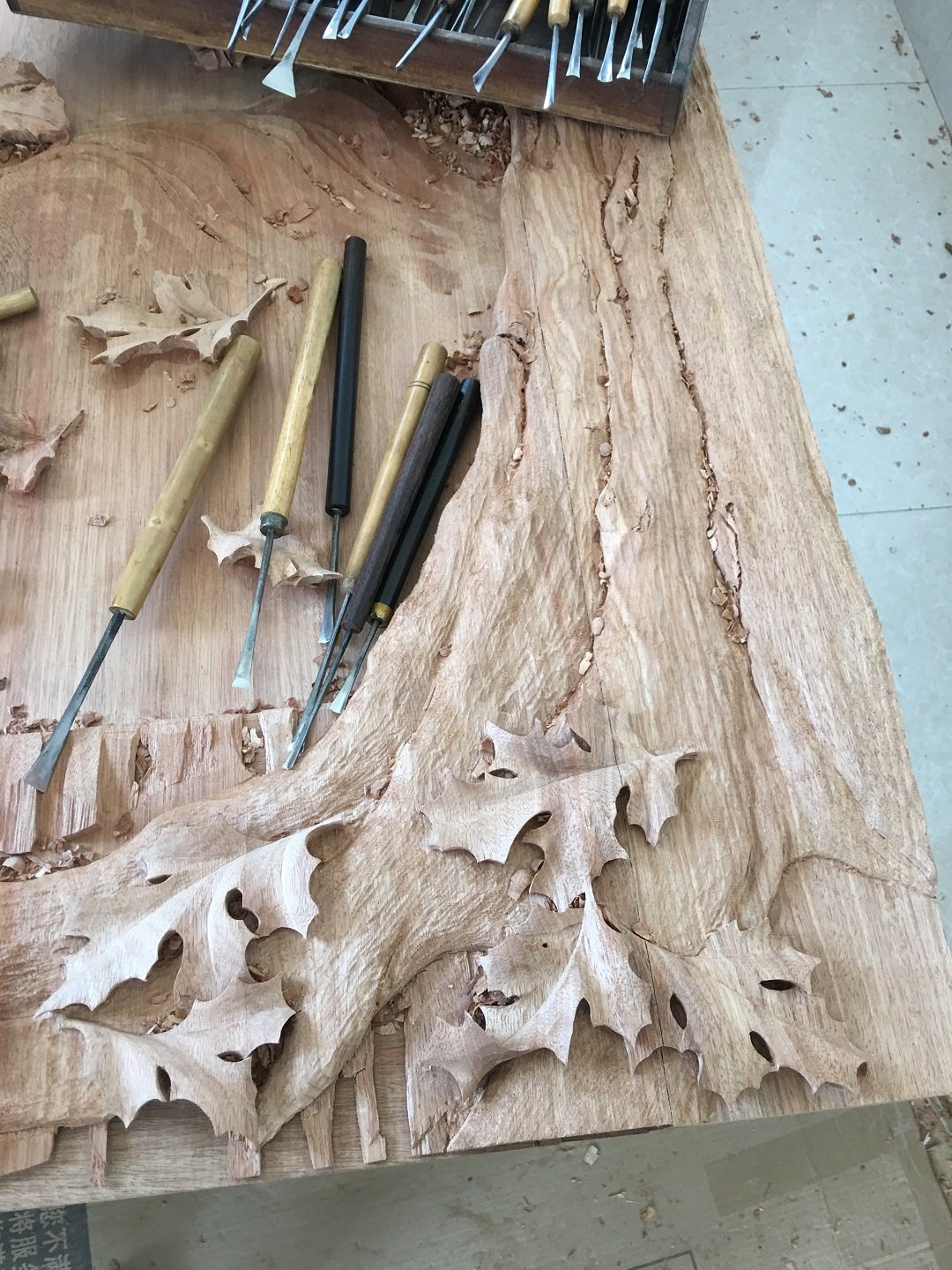 High-angle workshop view of hand-carving chisels resting on solid timber alongside deeply sculpted 3D oak leaves and organic tree trunk bark textures by Crown Custom Doors.