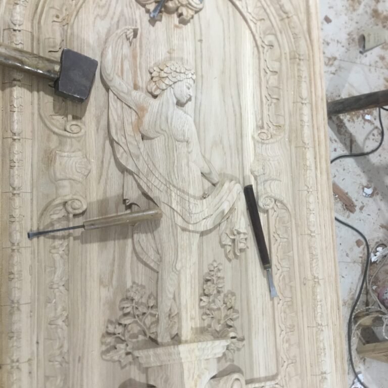 Overhead workshop view of a large custom solid wood door panel featuring a fully hand-carved classical female figure, arched moldings, and floral details, accompanied by traditional woodcarving chisels and a mallet.