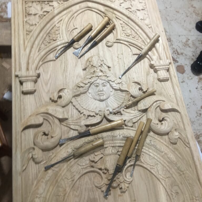 Overhead shot of a raw solid wood door panel featuring a hand-carved Gothic pointed arch tracery, a sculpted face with a ruff collar, and classical scrollwork, decorated with traditional woodcarving chisels at Crown Custom Doors.