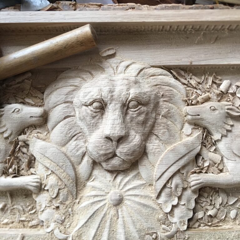 Extreme close-up of a raw solid wood door header featuring a hand-carved lion head flanked by heraldic creatures, with fresh wood shavings and a chisel handle visible.
