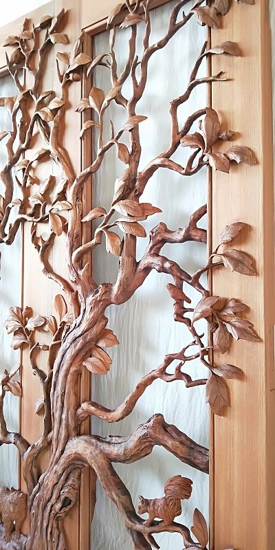 Angled close-up view of a handcrafted solid wood double door showcasing high-relief 3D carvings of an oak tree trunk, a squirrel, a songbird, and dense leaf canopy over glass.