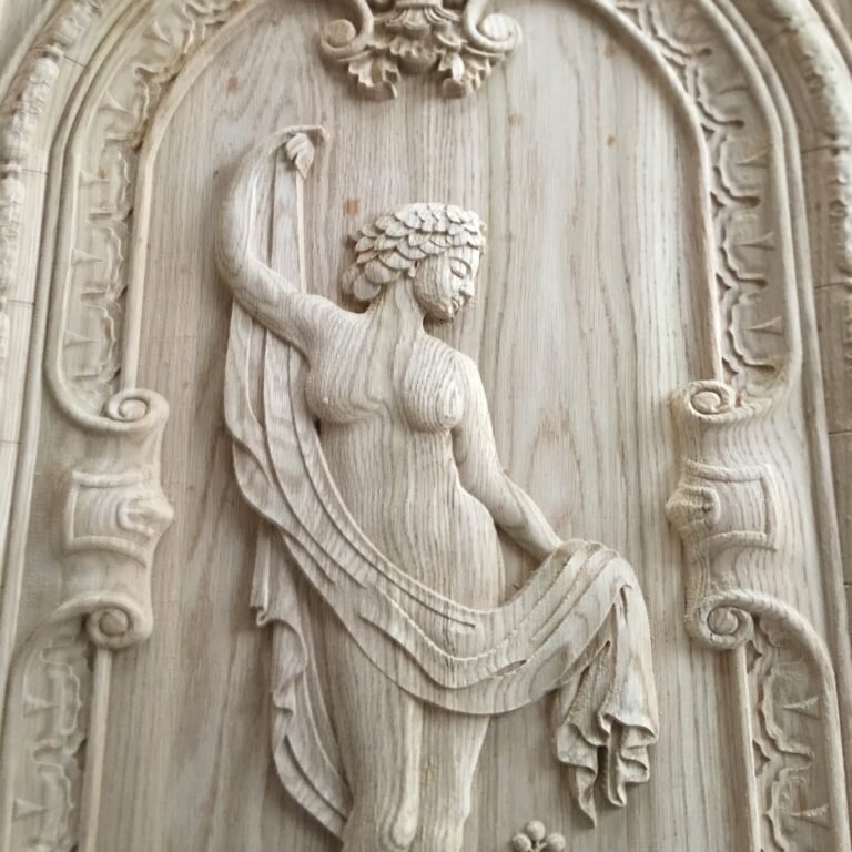 Extreme close-up of a custom raw solid wood door panel featuring an intricate hand-carved classical female figure draped in flowing fabric within an ornate arched frame with scrollwork details by Crown Custom Doors.