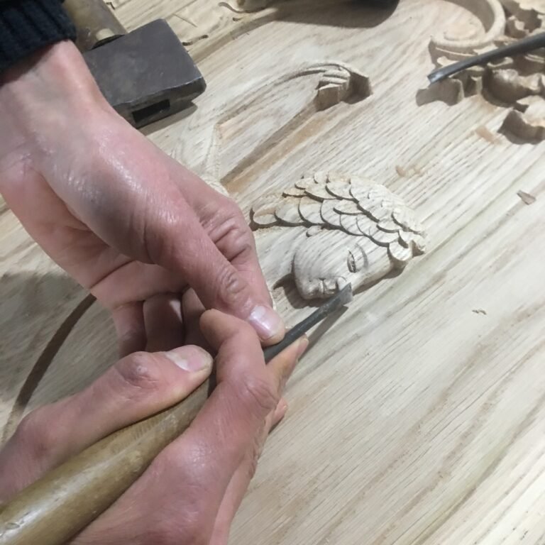 Extreme macro close-up of a woodcarver using a small chisel to sculpt delicate facial details and textured hair on a custom solid wood door panel at Crown Custom Doors.