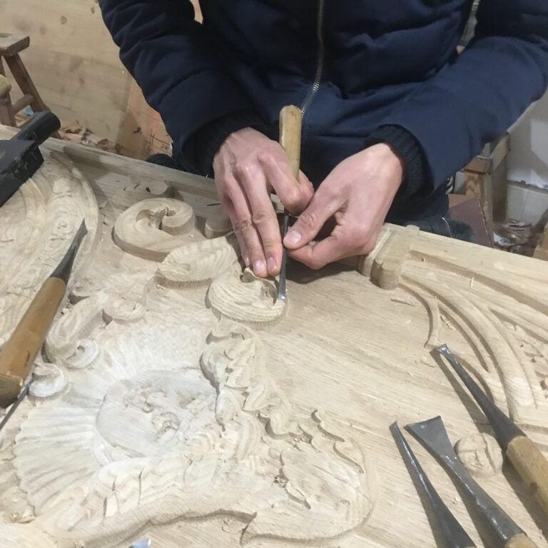 Extreme close-up shot of a woodcarver's hands precisely carving classical scrollwork motifs on a solid wood door panel using fine gouge chisels at Crown Custom Doors.