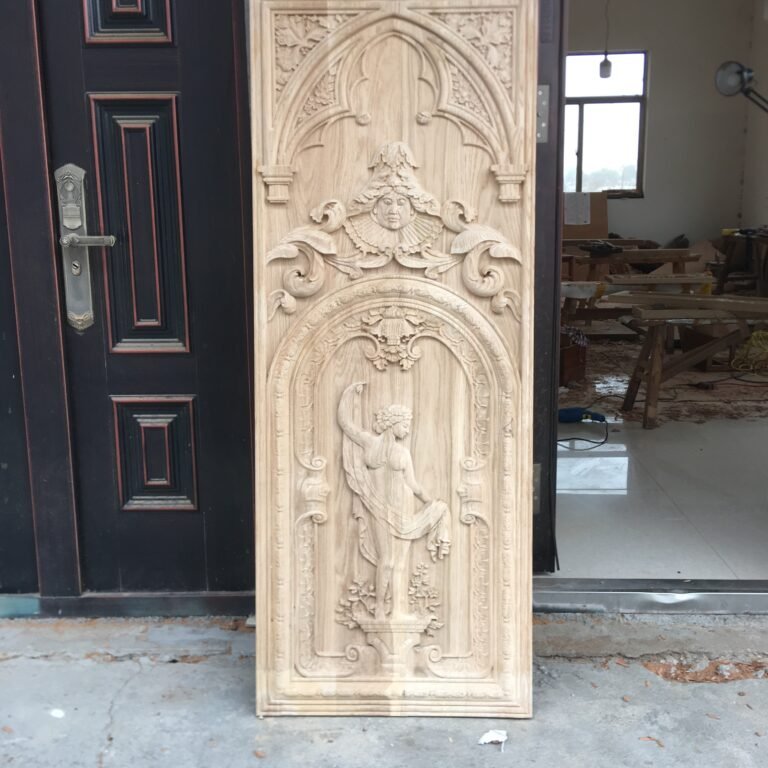 Full vertical shot of a raw, unfinished solid wood door panel featuring intricate hand-carved Gothic pointed arches, a central face relief with ruff collar, and a classical draped female sculpture, standing upright at a workshop.