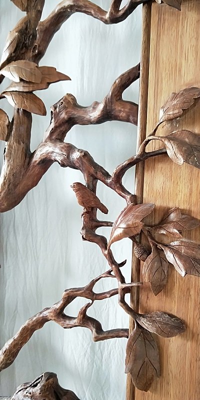 Close-up detail of a handcrafted solid wood door featuring a 3D relief carving of a songbird perched on an oak tree branch with carved oak leaves and an acorn accent.
