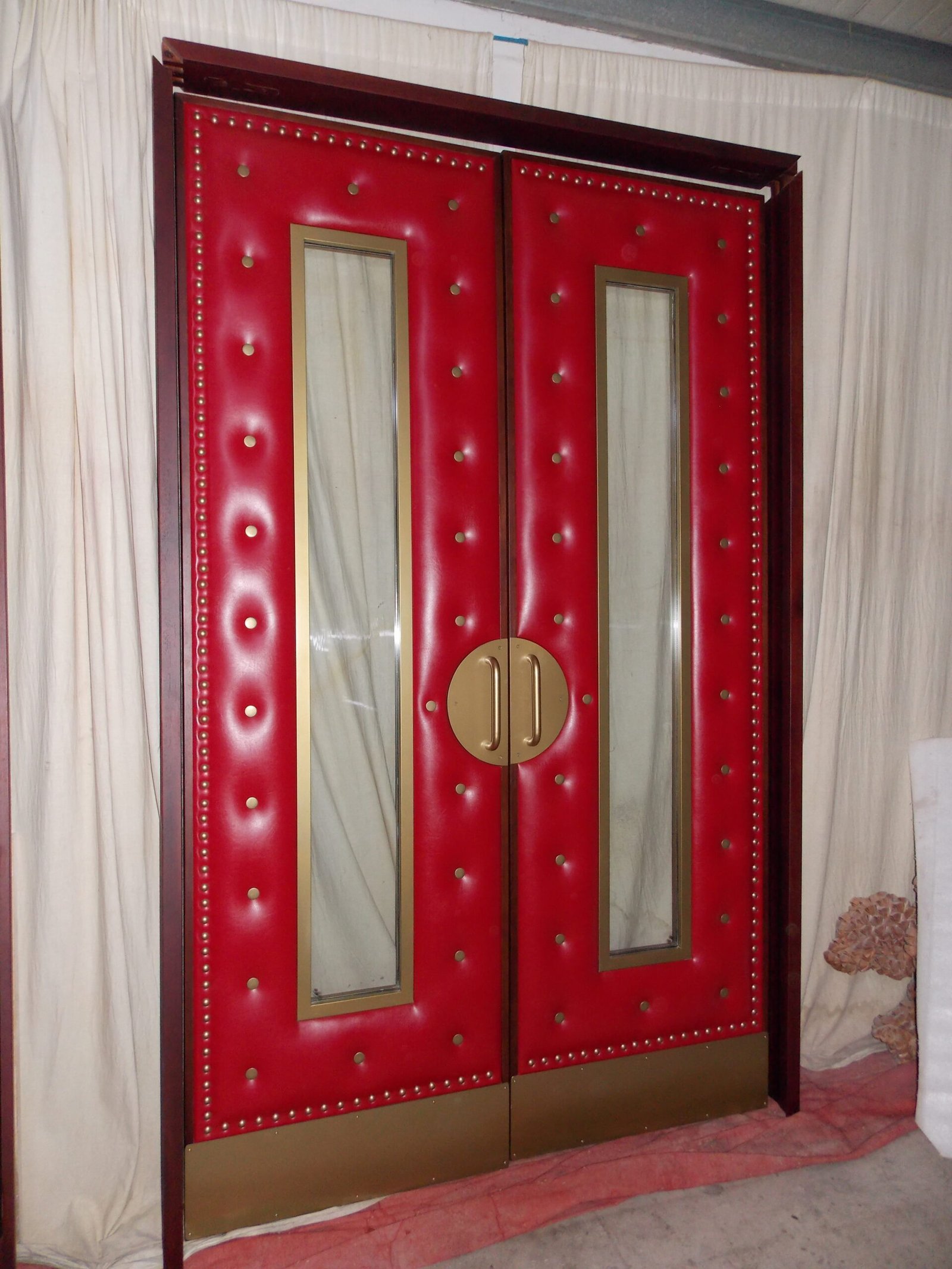 Full-length front view of handcrafted red upholstered soundproof double doors pre-hung in a mahogany wood jamb frame, featuring deep tufted vinyl leather, brass nailhead perimeter trim, gold-framed glass vision panels, and circular brass pull hardware.