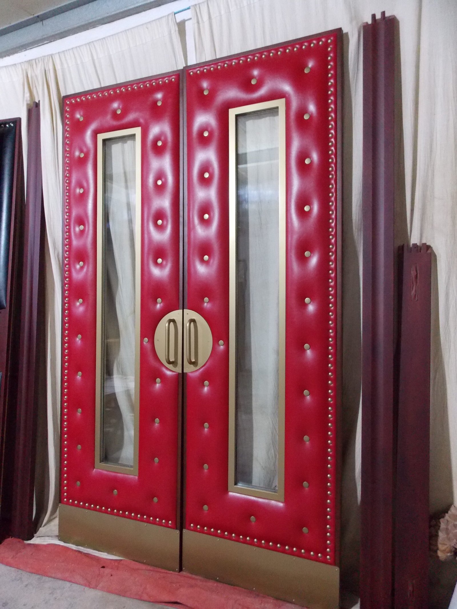 Custom soundproof home theater double doors with rich red tufted leather, brass nailhead trim, gold porthole glass panels, and solid brass pull handles.