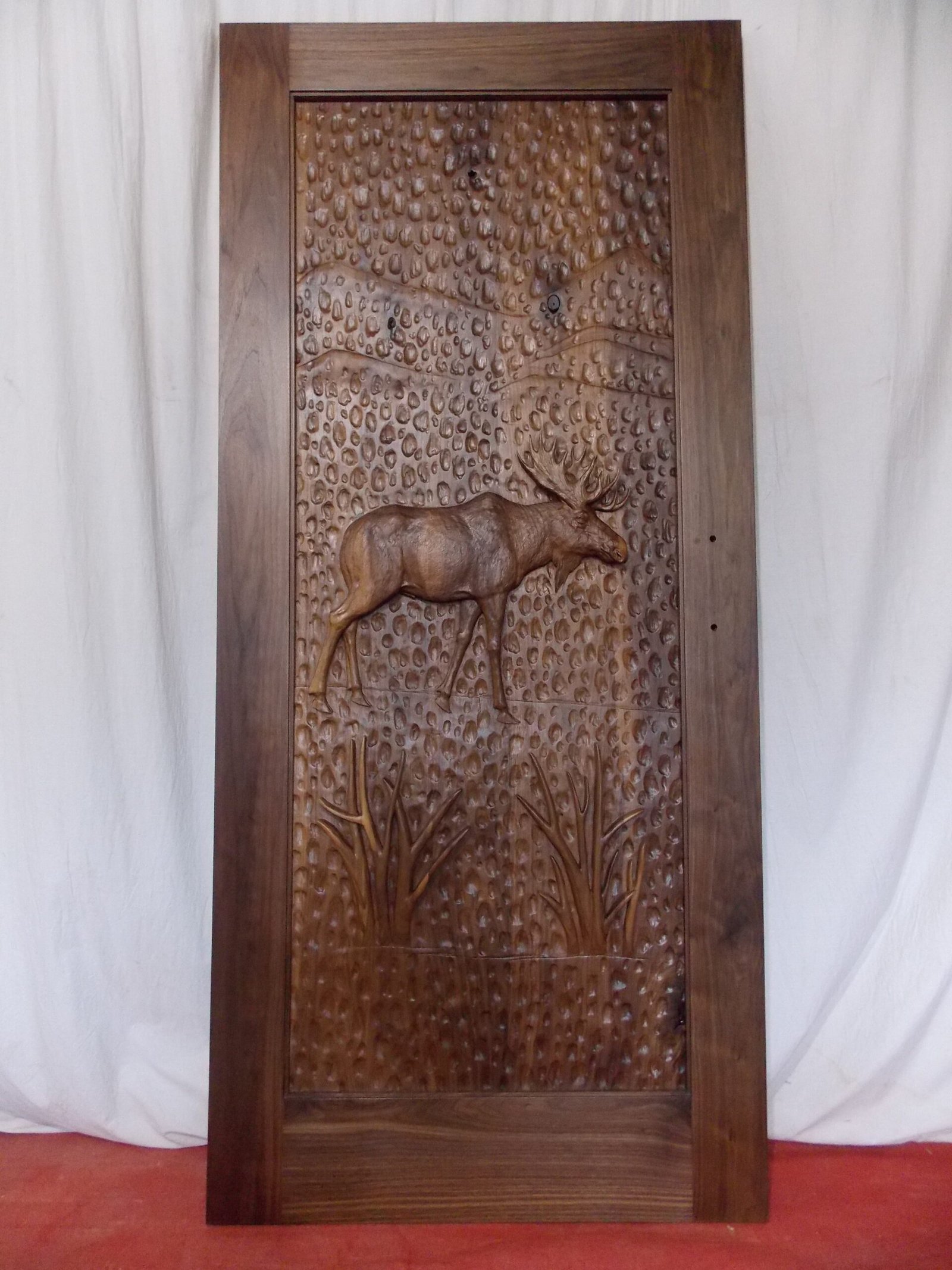 Full-length front view of a finished solid black walnut single entry door featuring a detailed high-relief hand-carved bull moose against a hand-gouged textured background, standing against a light studio backdrop.