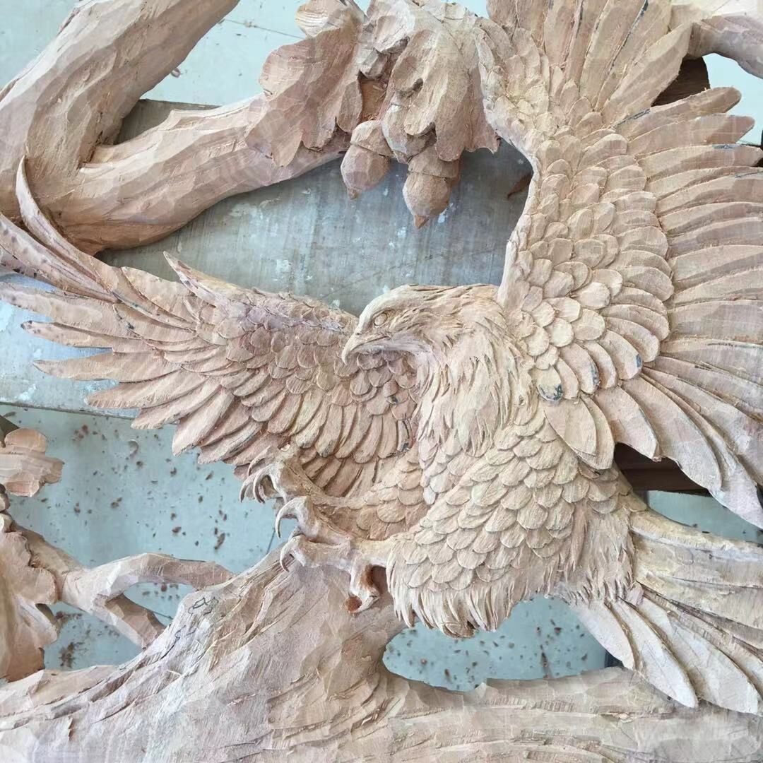 Workshop close-up view of a raw solid wood door panel featuring a high-relief hand-carving of an eagle in flight with outstretched wings and sharp talons reaching toward an oak tree branch with carved acorns.