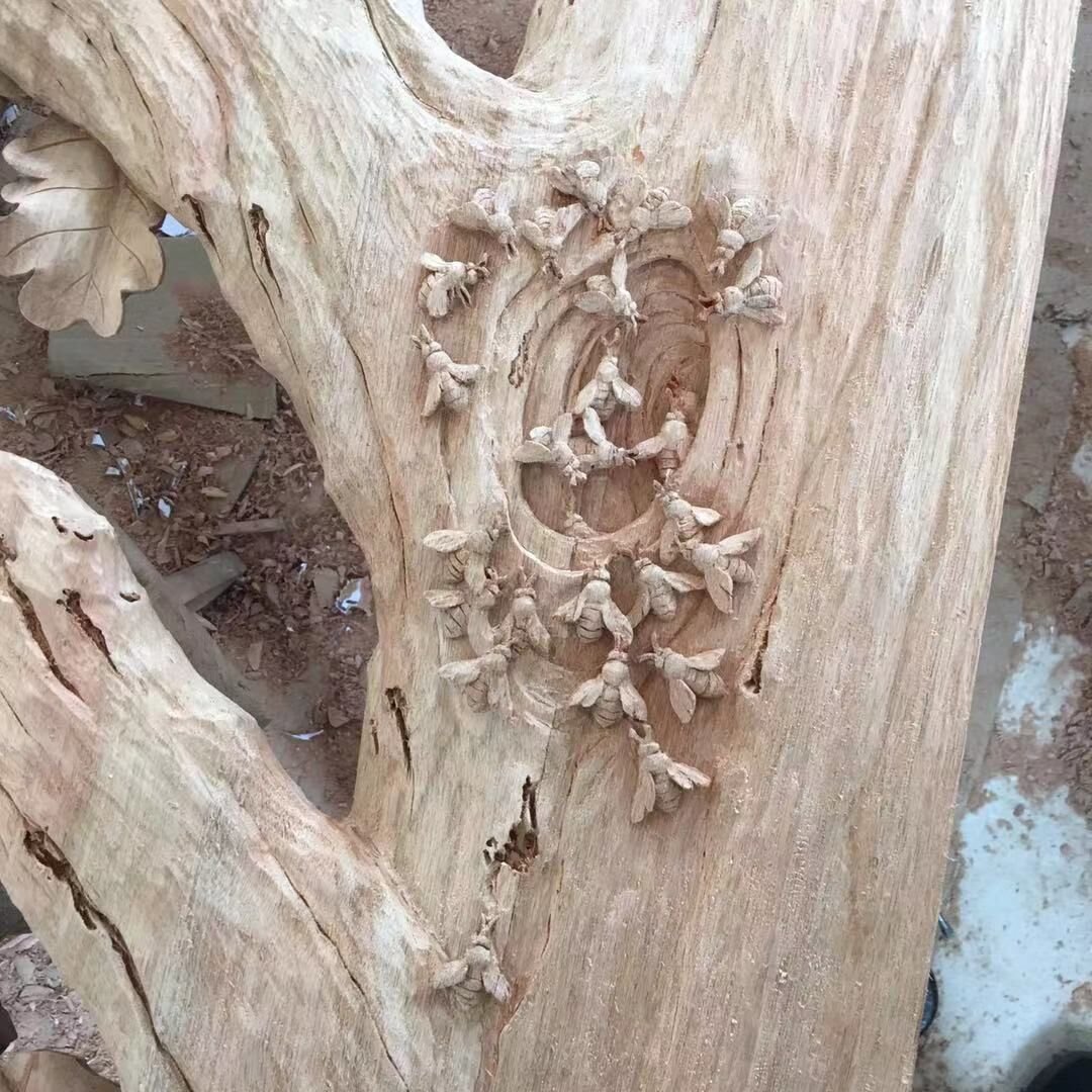 Workshop close-up of a raw solid wood door panel featuring a high-relief hand-carving of a swarm of honeybees crawling around a natural tree trunk hollow, with carved oak leaf contours nearby.