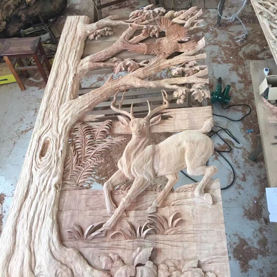 High-angle workshop view of a raw solid wood door panel featuring a high-relief hand-carved leaping deer framed by a large oak tree trunk, spreading canopy branches, ferns, and an eagle in flight.