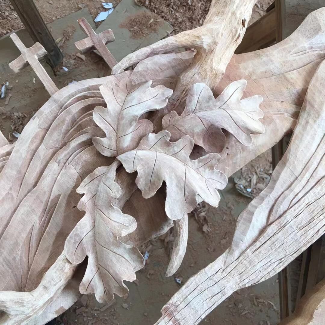 Workshop close-up of a raw solid wood door panel featuring high-relief carved oak leaves with delicate veining, sculpted tree branches, and small wooden crosses in the background.