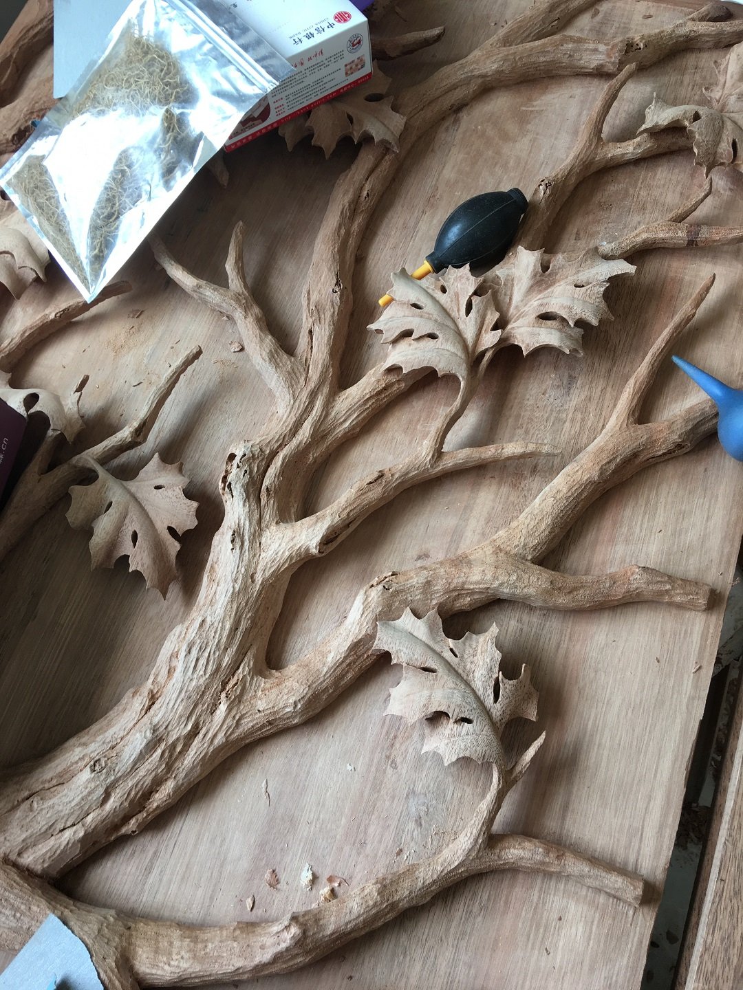Top-down workshop view of raw hand-carved solid wood tree branches and oak leaves with cleaning tools, showing 3D relief woodworking details by Crown Custom Doors.