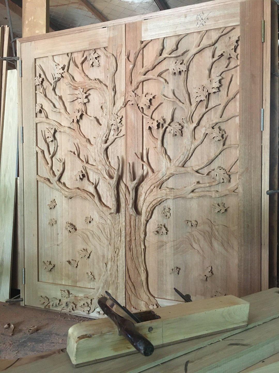 Pre-hung custom solid wood double entry door featuring an unvarnished hand-carved 3D oak tree relief motif with wooden hand plane tools resting in the foreground at Crown Custom Doors.