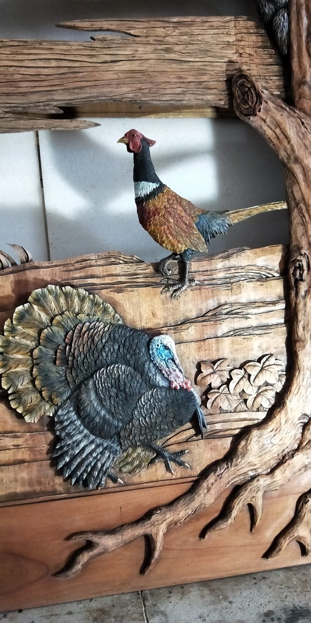 Close-up shot of a solid wood door panel featuring high-relief, hand-carved, and painted wildlife artwork of a strutting wild turkey with fanned tail feathers alongside a perched ring-necked pheasant set within a sculpted tree branch frame.