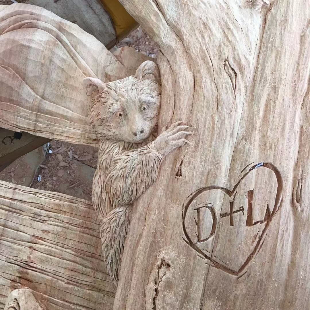 Close-up workshop view of a raw solid wood door carving showing a high-relief raccoon peeking out from behind a textured tree trunk with custom initials carved inside a heart.
