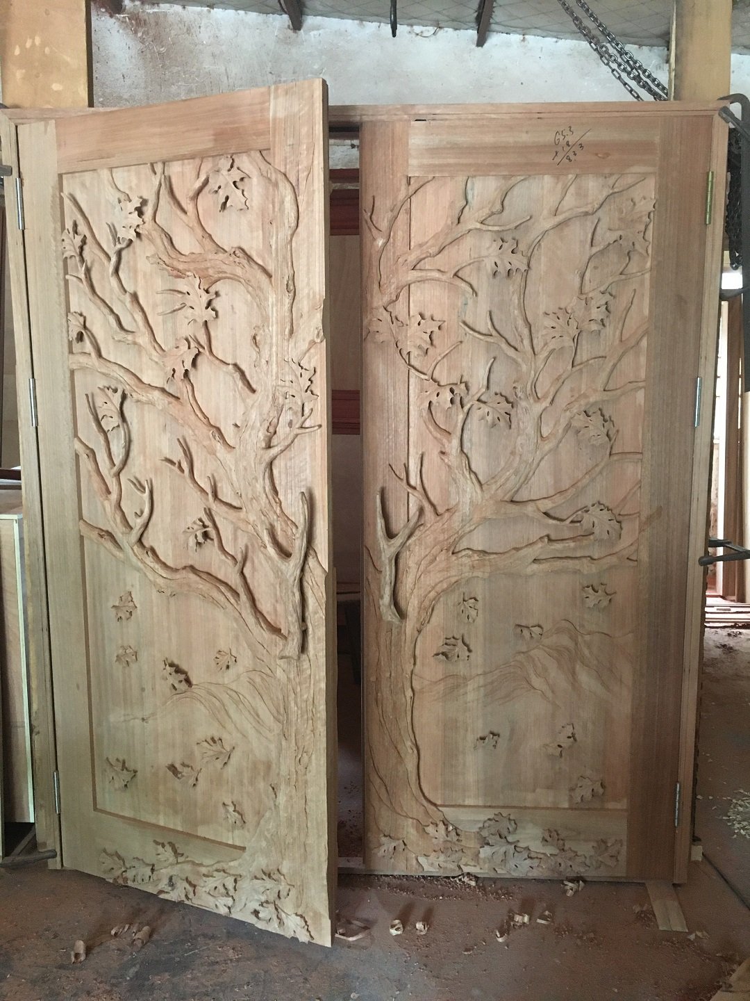 Custom handcrafted raw solid wood double entry door featuring an unvarnished 3D relief carving of a continuous oak tree with branches and falling leaves during frame assembly in the Crown Custom Doors workshop.