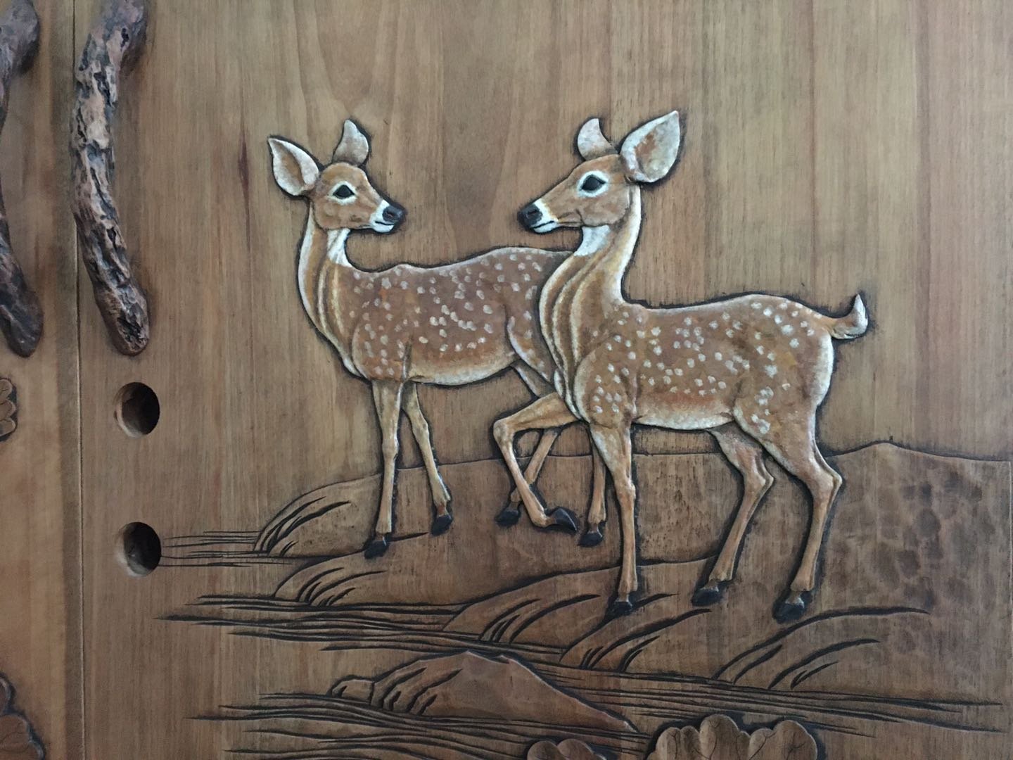 Close-up of hand-carved and painted spotted deer fawns relief on a solid wood door panel featuring natural wood branch handles, crafted for a rustic log cabin or mountain lodge home.