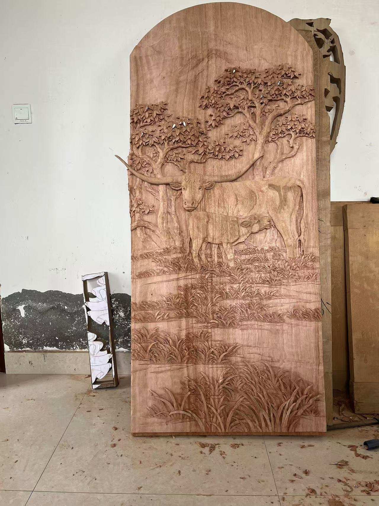 Full-length vertical view of an unfinished hand-carved arched solid wood door panel standing upright in a workshop, featuring a high-relief Texas Longhorn cow and calf for a rustic ranch home.